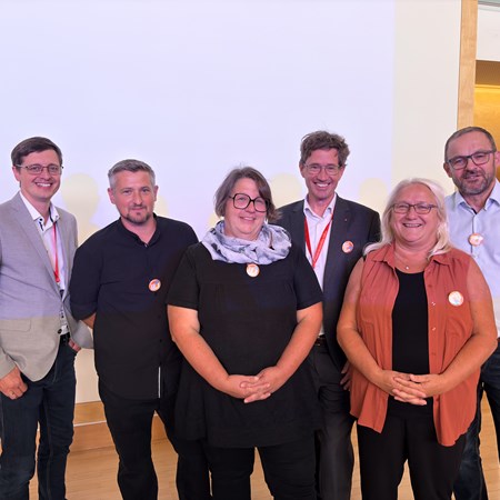 Sechs Personen stehen lächelnd nebeneinander, einige mit Ansteckern, im Innenraum.