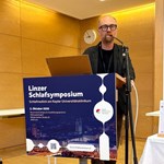 Linzer Symposium: Mann spricht am Rednerpult vor Logo der Kepler Universitätsbibliothek