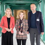 Drei Personen stehen in einem Gang, links eine Frau im roten Blazer, Mitte eine Frau mit kariertem Blazer, rechts ein Mann mit dunklem Sakko.