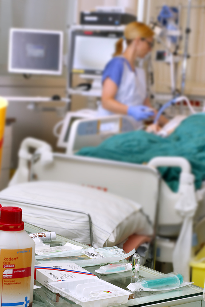 Medikamente auf einem Tisch, dahinter Patientenbett auf Intensivstation mit Pflegekraft